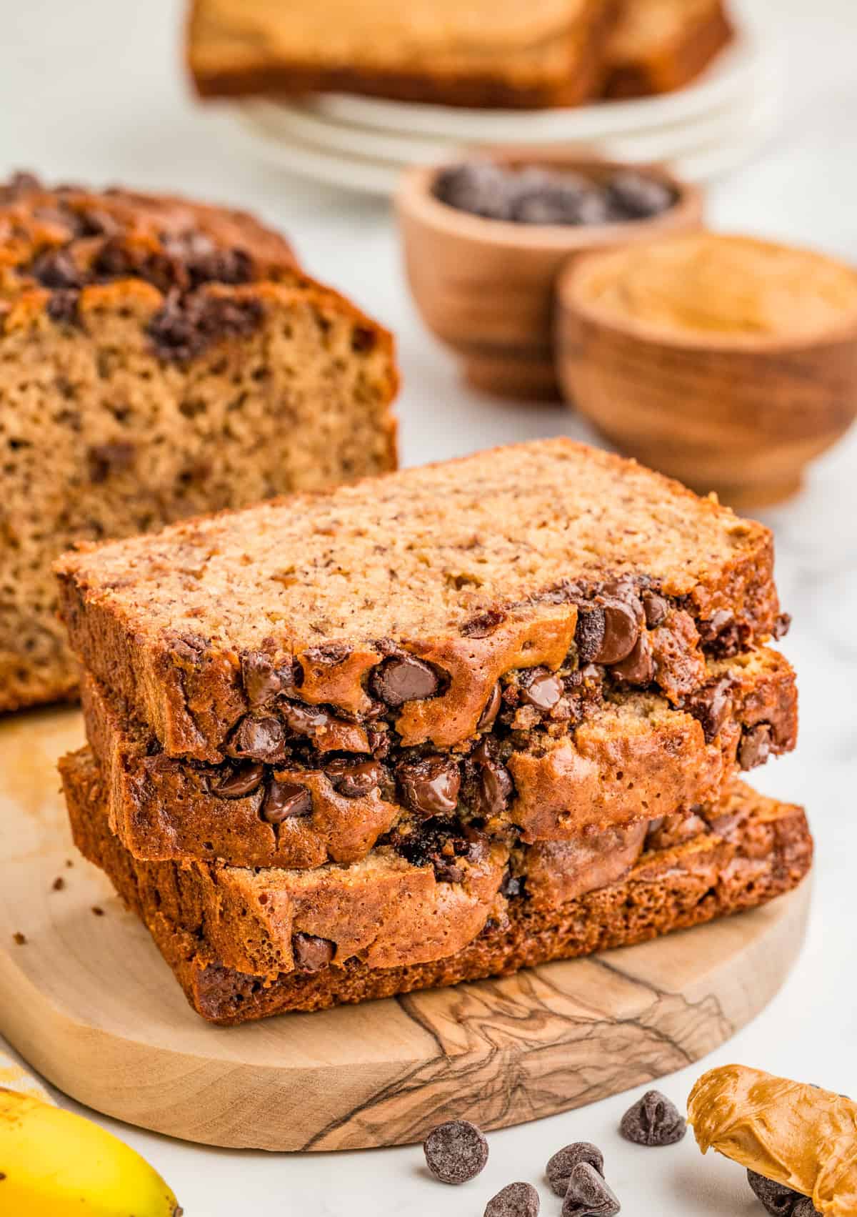 Three stacked slices of Peanut Butter Banana Bread on wooden board with peanut butter and chocolate chips in background.