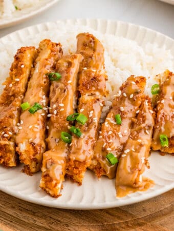 Close up square image of cut up chicken over rice on white plate with garnishes.