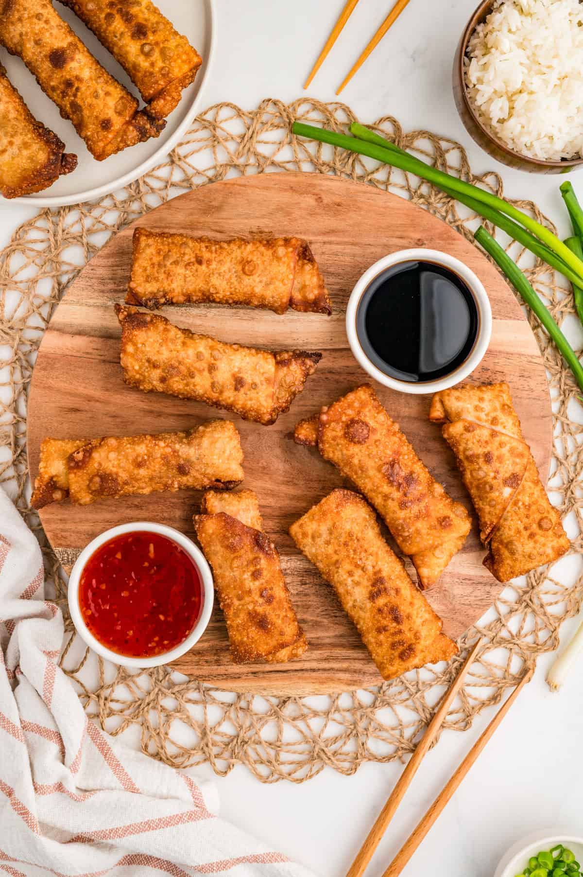 Overhead image of egg rolls on wooden board with sweet and sour sauce, soy sauce and chopsticks.