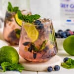 Square image of cocktail on marble coaster with limes and blueberries around it.