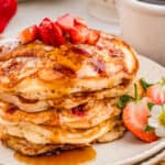 Close up square image of pancakes stacked on plate topped with powdered sugar, syrup and strawberries.