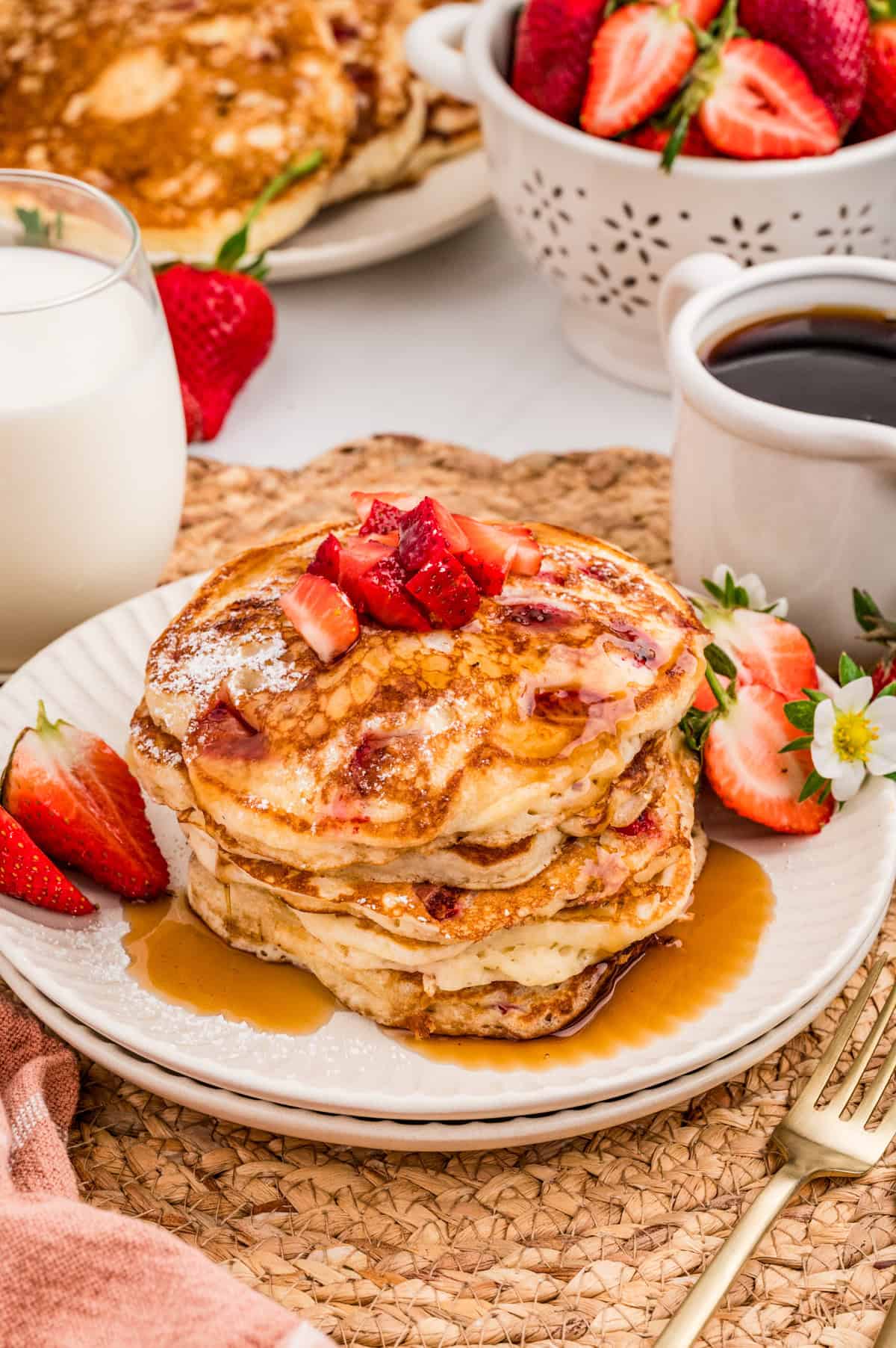 Finished pancakes on stacked plates topped with strawberries, syrup and powdered sugar. Coffee, milk and strawberries behind plate.