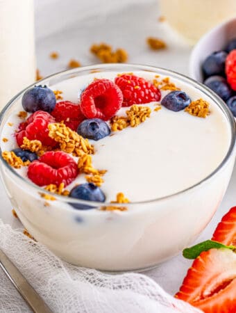 Square image of yogurt in a clear bowl topped with granola and berries.