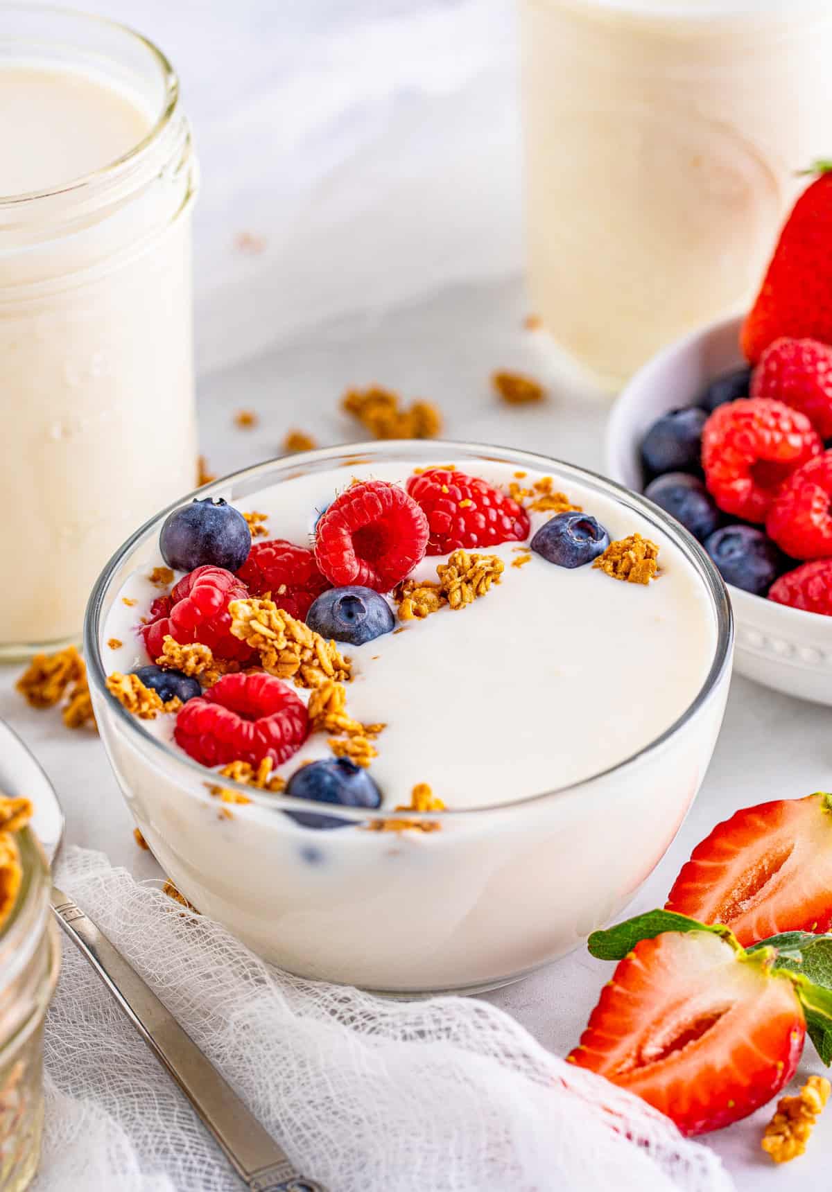 Instant Pot Yogurt in a clear bowl topped with granola and berries with sliced strawberries in front and yogurt in jars behind.