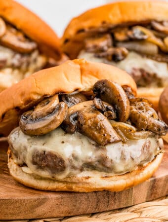 Square image of burgers on wooden board with mushrooms and melted cheese.