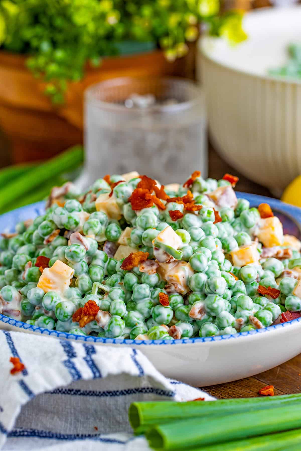 Pea Salad in serving bowl topped with bacon with green onions in front of bowl and a drink behind.