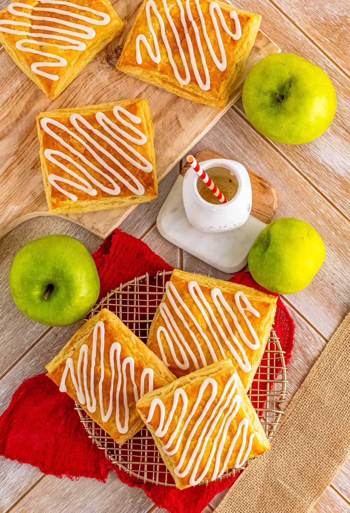 Overhead of finished strudel on wire rack and wooden board with milk and apples around them.
