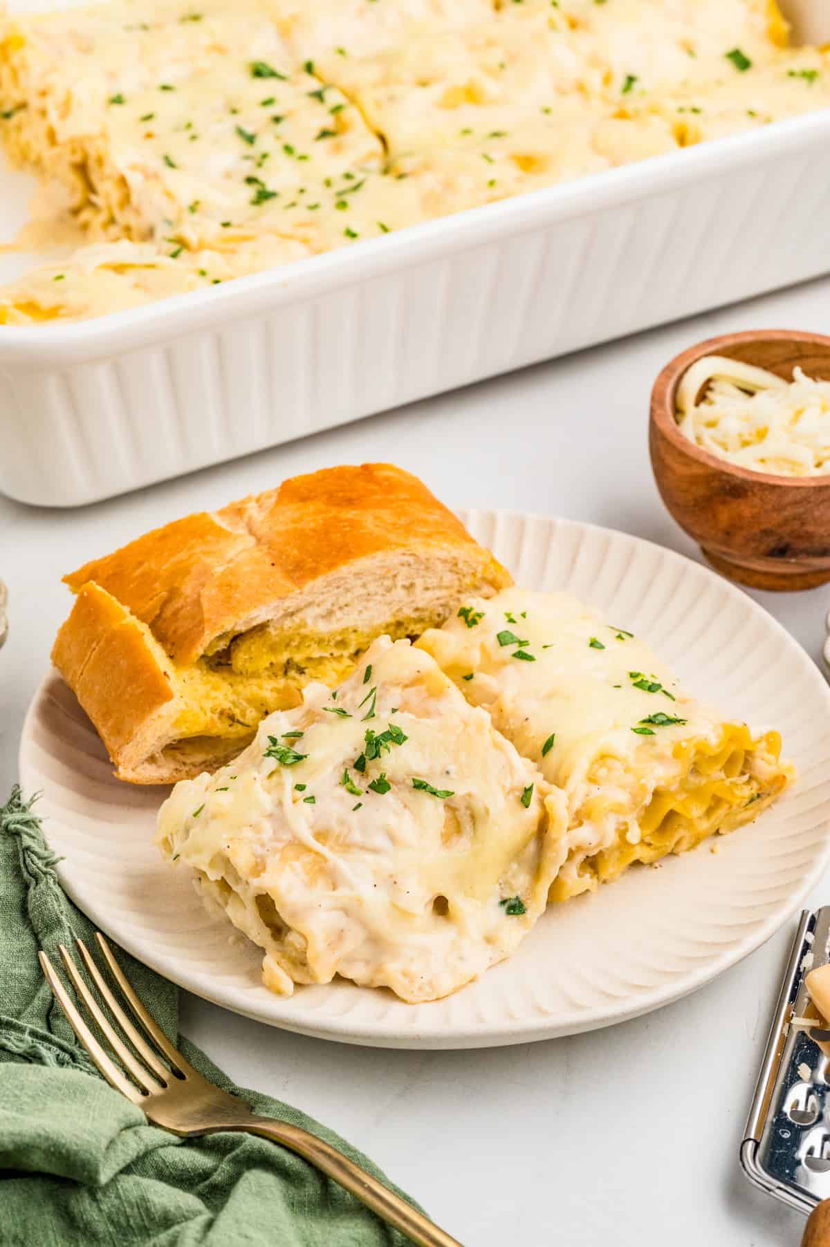 Two Chicken Alfredo Lasagna Rolls on plate with garlic bread. Pan of rolls behind the plate.