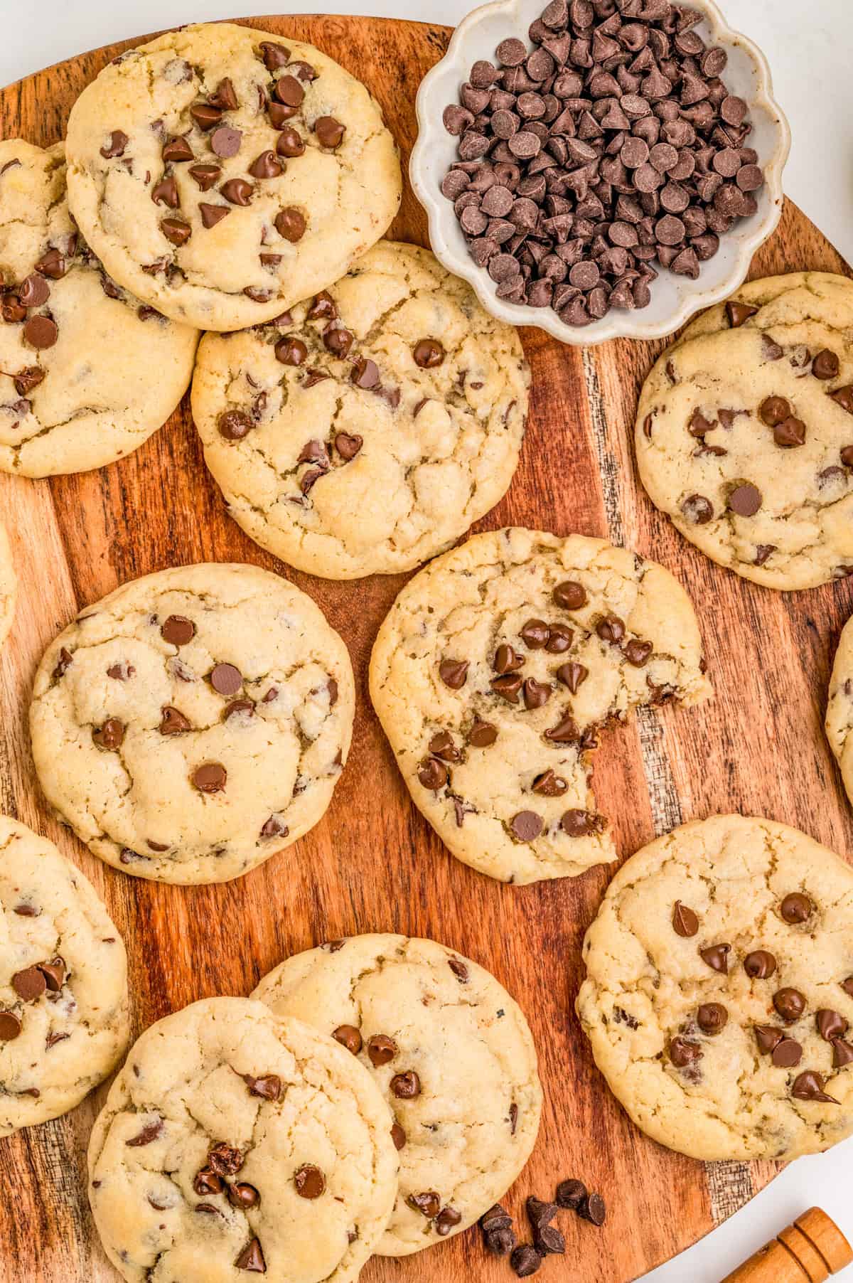 Overhead of Cream Cheese Chocolate Chip Cookies on wooden board spread out one with bite taken out and a bowl of chocolate chips.