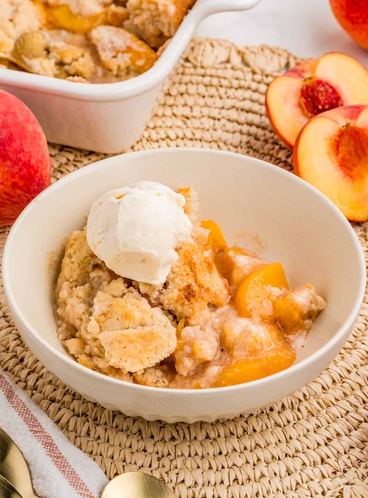 Peach Cobbler in bowl with a scoop of ice cream. Pan of cobbler and peaches in background.