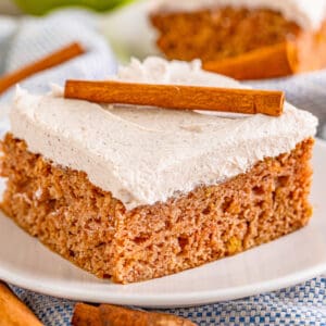 Close up square image of cake on white plate frosted and topped with a cinnamon stick.
