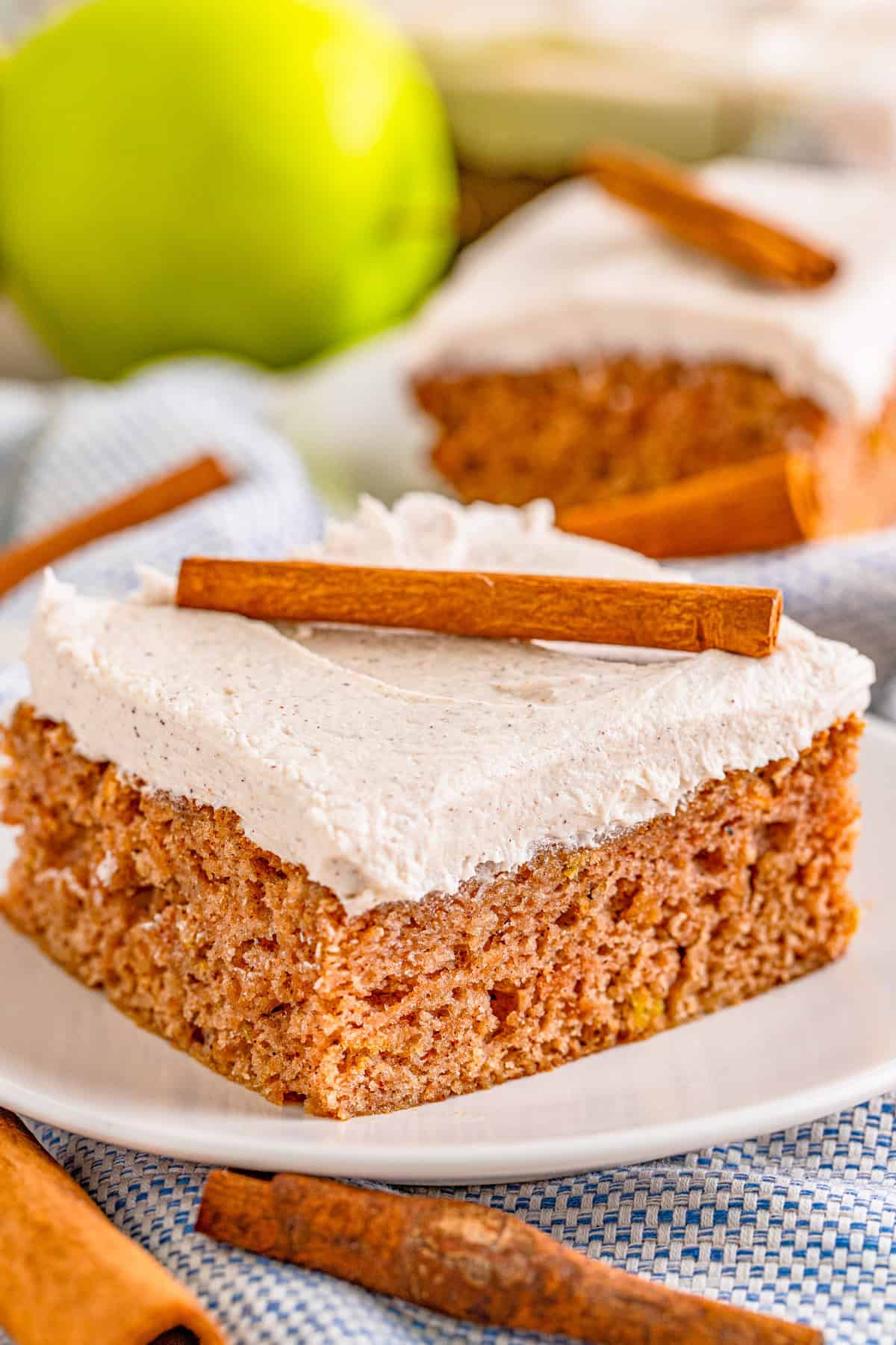 Slice of Apple Cake topped with frosting and a cinnamon stick with a slice of cake in background and apple.