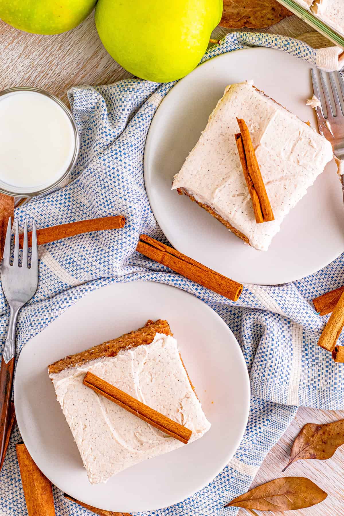 Overhead photo of Apple Cake slices on white plates topped with frosting and cinnamon sticks.