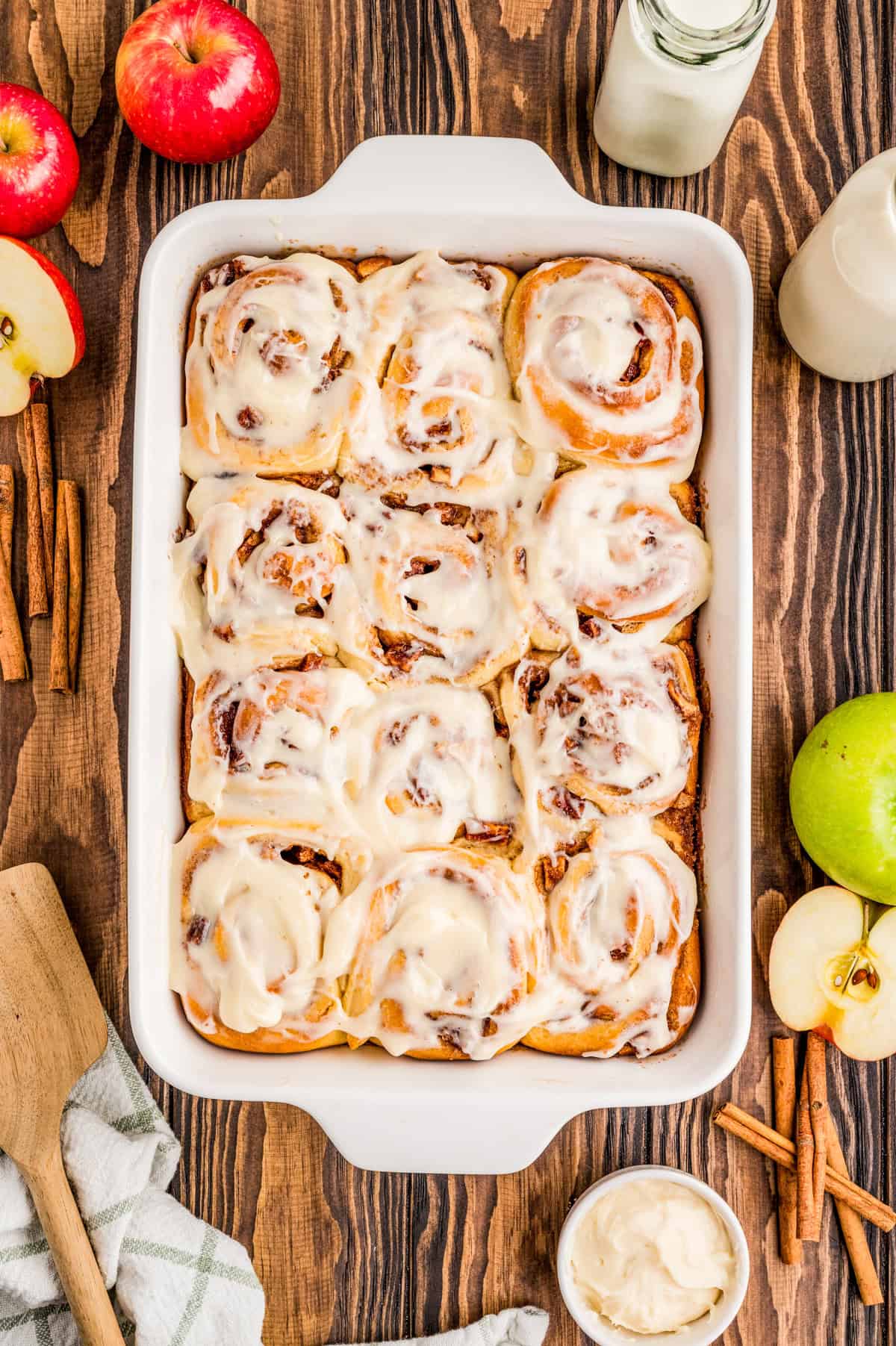 Overhead of finished frosted cinnamon rolls surrounded by apples and cinnamon sticks.