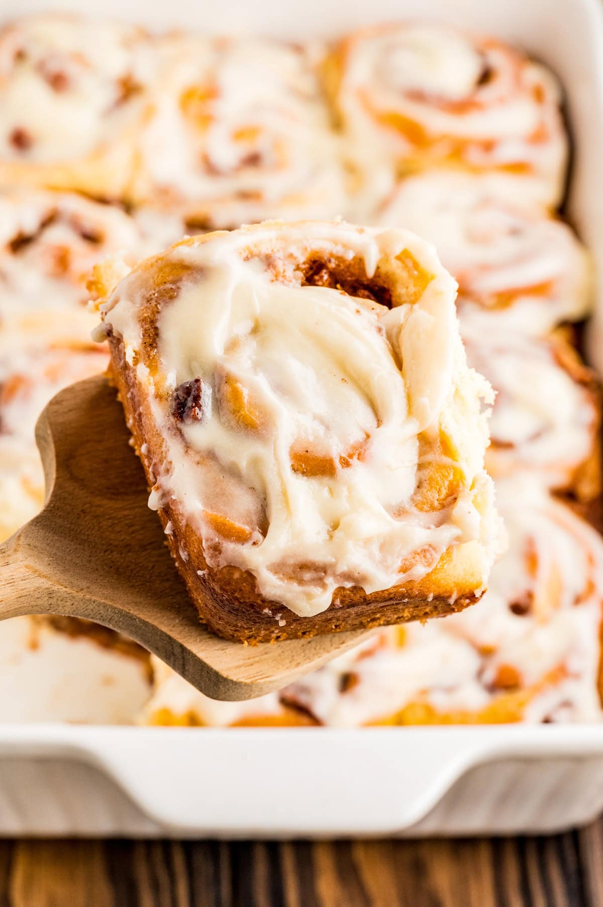 Wooden spatula holding up one of the cinnamon rolls out of the pan.