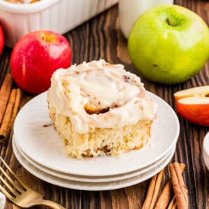 Square image of one of the cinnamon rolls on stacked white plates with apples and cinnamon by plate.
