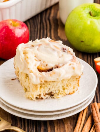 Square image of one of the cinnamon rolls on stacked white plates with apples and cinnamon by plate.