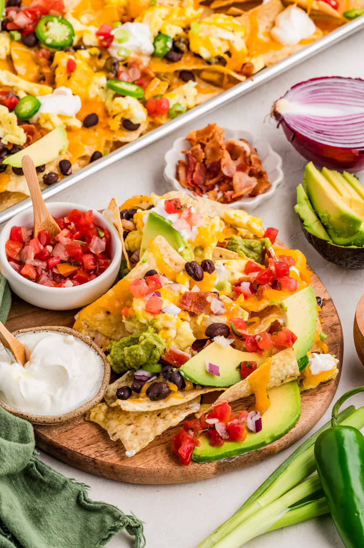 Breakfast Nachos on wooded round board with pico and sour cream with full pan of nachos in background.