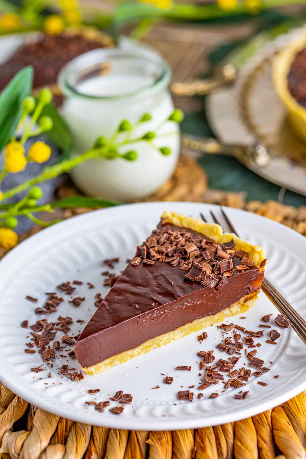 Slice of the Chocolate Ganache Tart on white plate with chocolate curls and fork with milk in background.