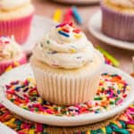 Close up square image of frosting on a cupcake on a white plate filled with rainbow sprinkles.
