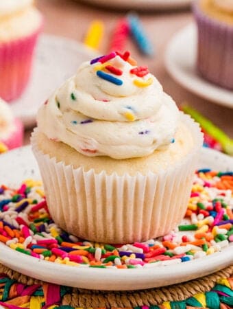 Close up square image of frosting on a cupcake on a white plate filled with rainbow sprinkles.