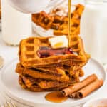 Square image of stacked waffles on white plate with cinnamon sticks with syrup being poured over the stack.