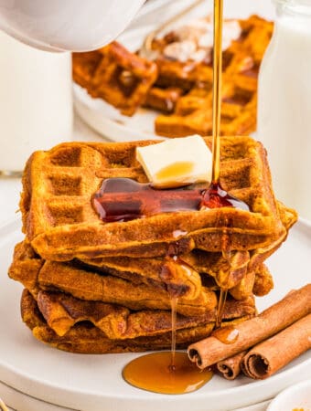 Square image of stacked waffles on white plate with cinnamon sticks with syrup being poured over the stack.
