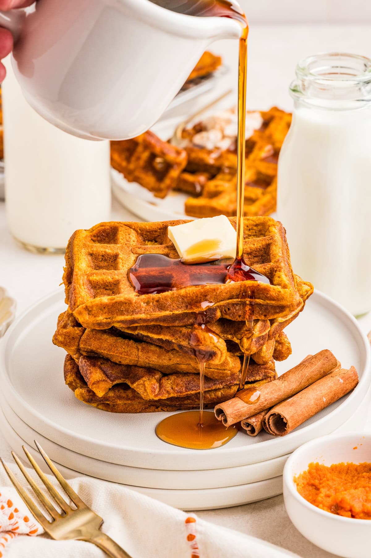 Stacked Pumpkin Waffles on white plate with cinnamon sticks and syrup being poured over stack.
