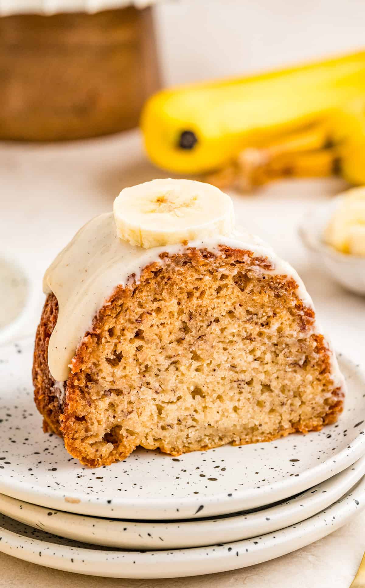Slice of Banana Bundt Cake frosted and topped with a slice of banana on white plate with bananas in background.