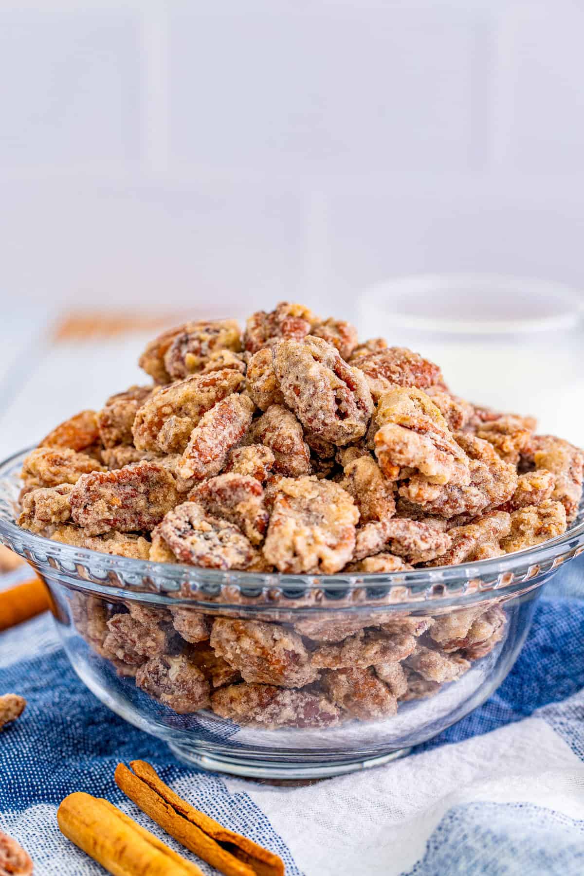Pecans in clear bowl with blue linen with cinnamon sticks in front of it.