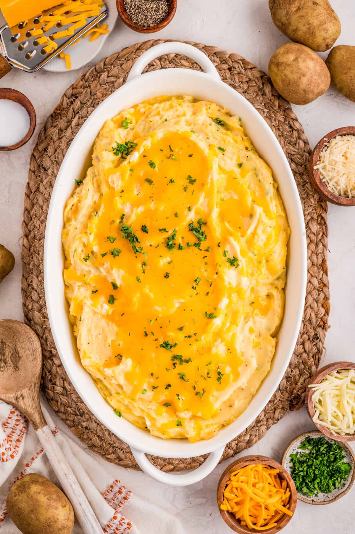 Overhead photo of finished Cheesy Mashed Potatoes in baking dish topped with cheese and parsley with other ingredients around dish.