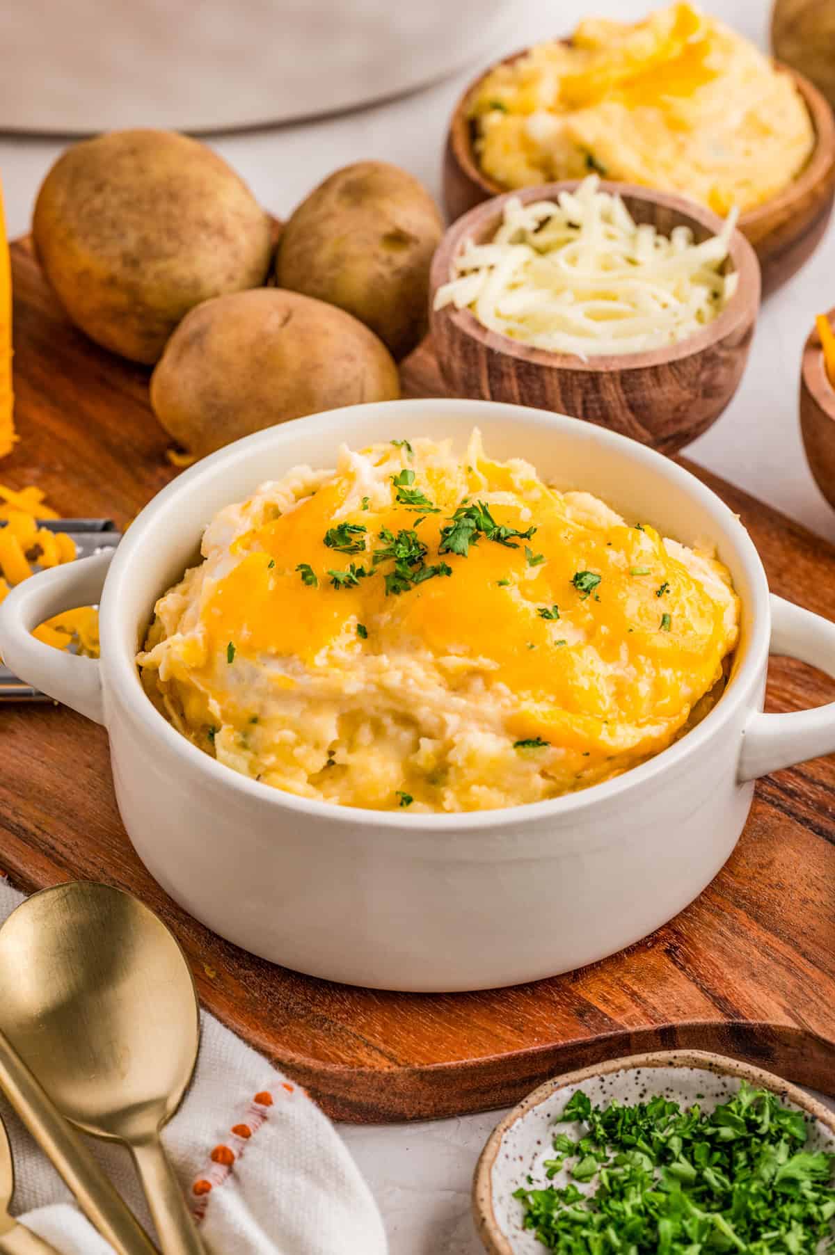Finished Cheesy Mashed Potatoes in a white handled bowl topped with parsley on a wooden cutting board with ingredients around bowl.