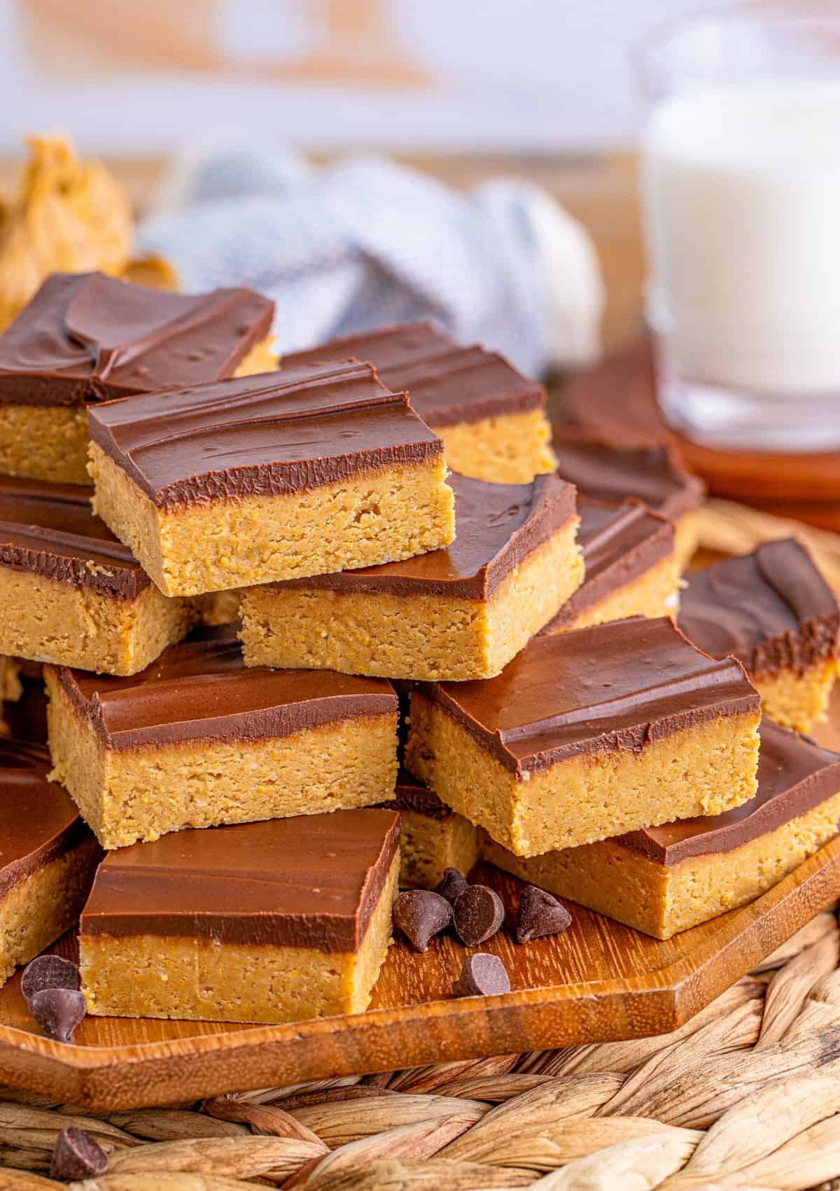 No Bake Peanut Butter Bars stacked on wooden platter with chocolate chips on platter and milk in background.