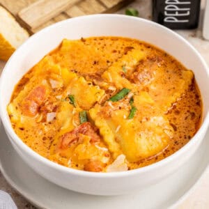 Close up square image of finished soup in white bowl on plate topped with basil and cheese.