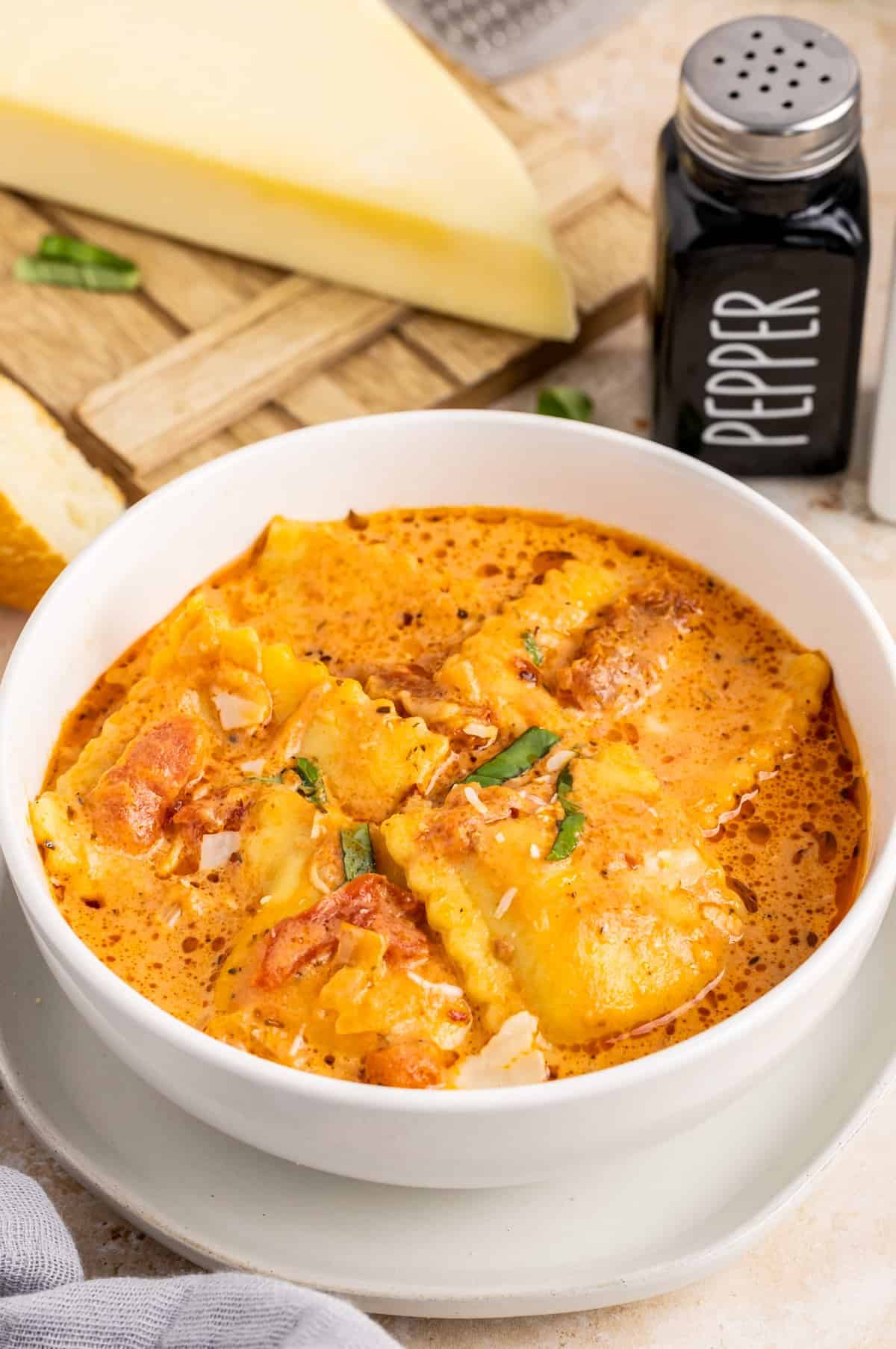 Ravioli Soup in white bowl on white plate topped with basil and cheese.