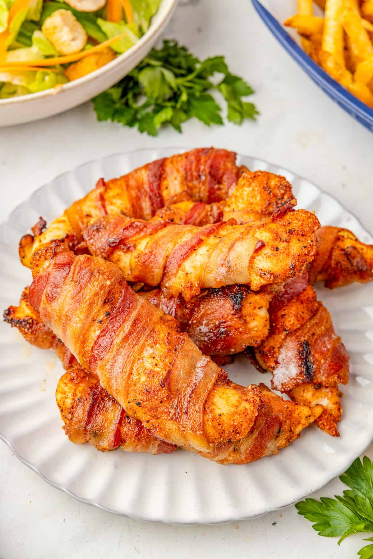 Bacon Wrapped Chicken Tenders stacked on top of one another on white plate with side dishes behind the plate.